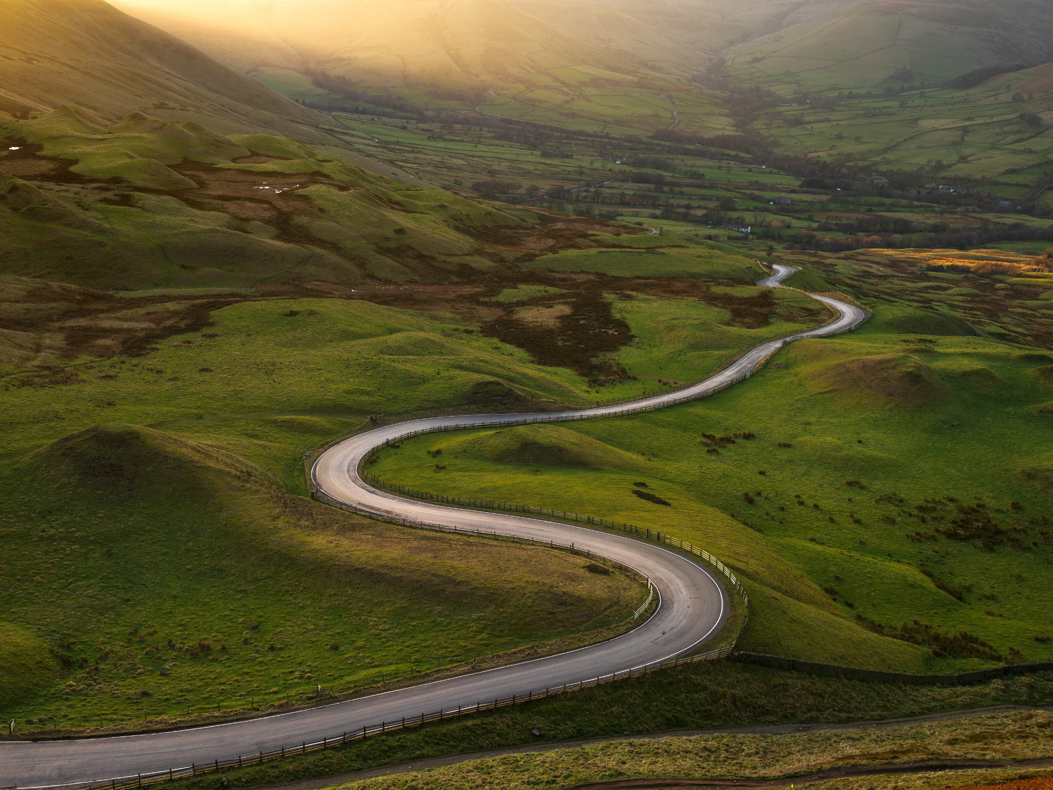 Winding road though green fields with the sun light pouring over the mountains 
